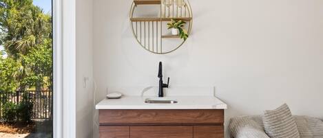 A bathroom with a white vanity, a black faucet, a wooden cabinet, a round mirror with a plant, and a view of trees outside.