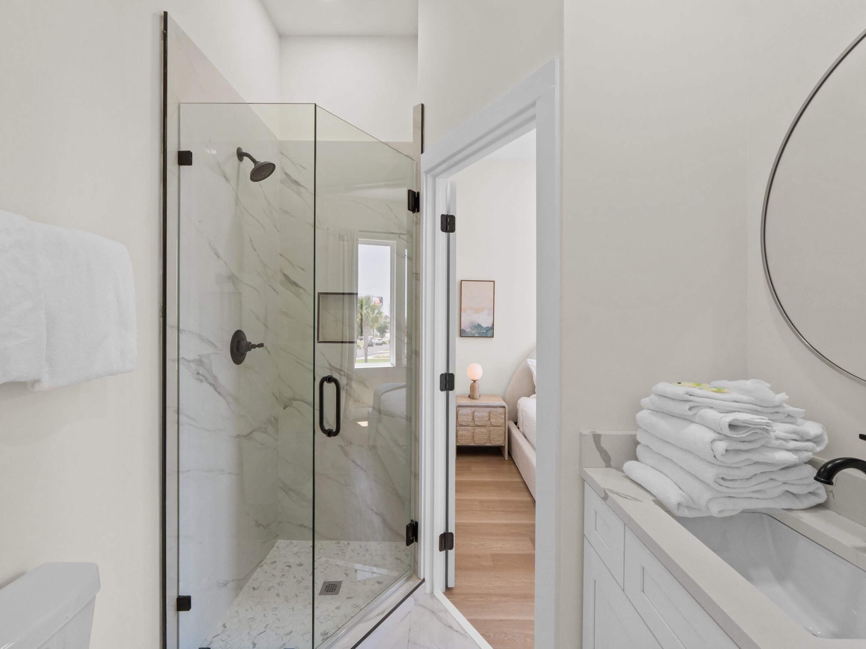 A modern bathroom with a glass shower enclosure, a sink with stacked towels, and a view into a bedroom.