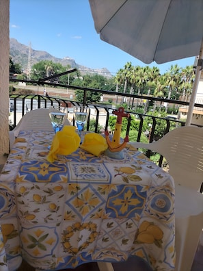 Un tavolo con tovaglia fantasia, un bicchiere d'acqua, un bicchiere di vino e un oggetto decorativo su un balcone con vista su una strada e del verde.