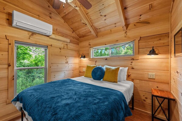 A wooden cabin bedroom with a bed, two windows, an air conditioner, and a ceiling fan. - New River Views - Cozy Cabin with Fire Pit (Todd)