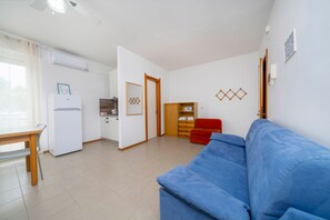 Ein Wohnbereich mit blauem Sofa, einem Holztisch, einer Küche mit Kühlschrank und einem Holzschrank.