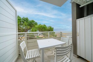 Ein Balkon mit weißem Tisch und Stühlen, der Blick auf einen Strand mit Bäumen und einem klaren Himmel bietet.