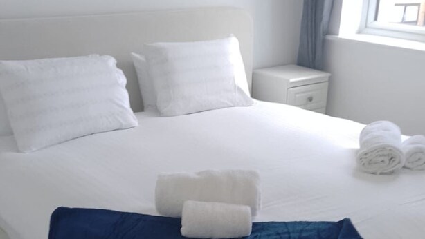 A neatly made bed with white linens and a blue throw blanket, a white nightstand with a drawer, and a window with curtains. - Huge 4 Bed House with parking and garden! (Bristol)