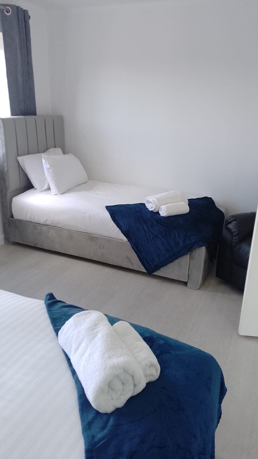 A single bed with white bedding and a blue blanket, a pillow, and a rolled towel on the bed.