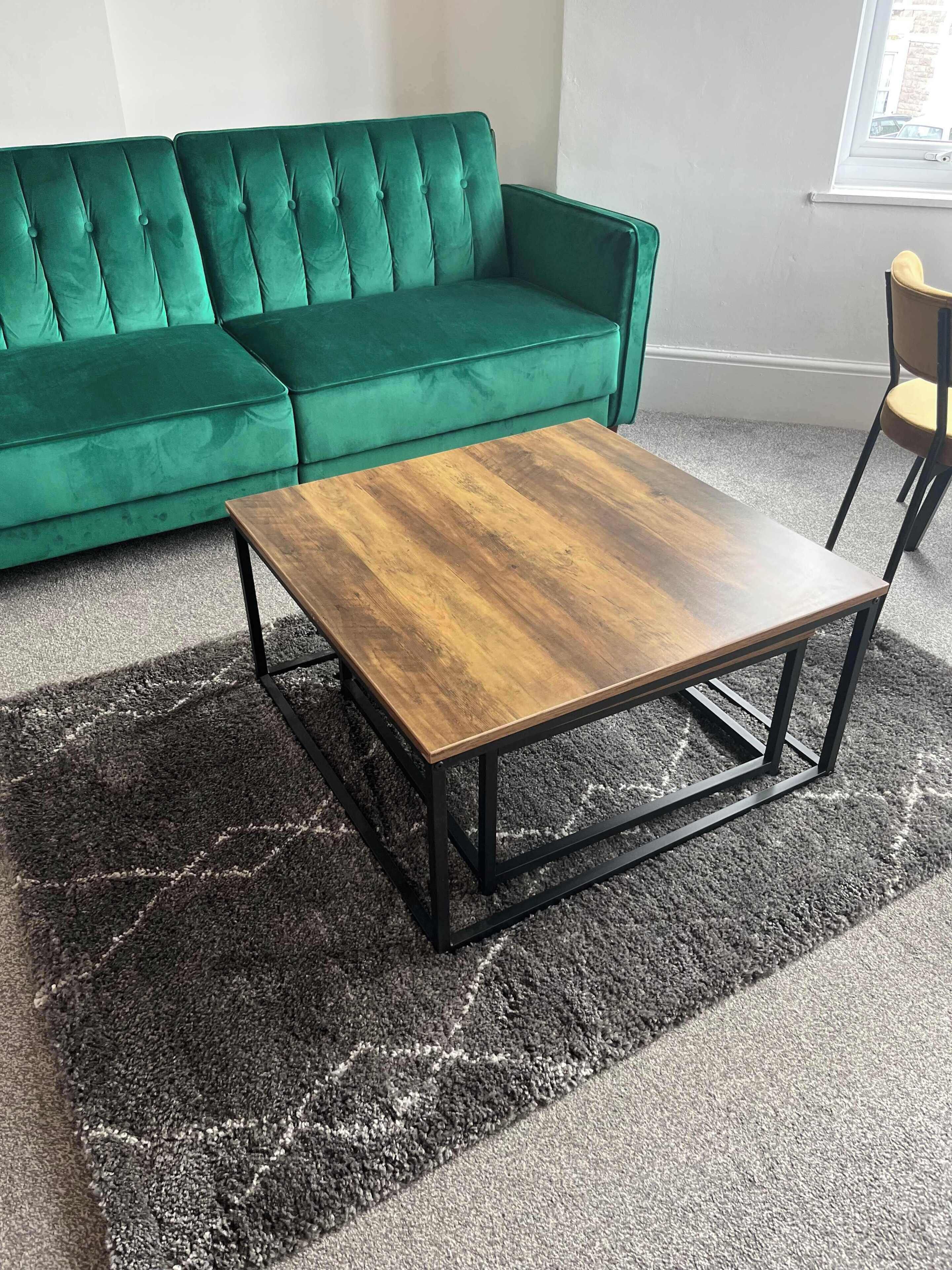 Un canapé vert, une table basse en bois avec des pieds en métal et un tapis à motifs.