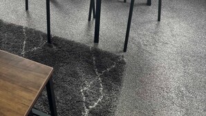 Un tavolo con piano in vetro, quattro sedie, pavimento rivestito di moquette e una finestra con vista su edifici e auto parcheggiate.