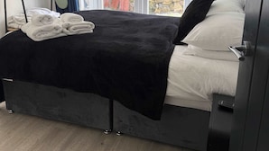 A bedroom with a bed, a window with curtains, a bedside table, and a wall-mounted heater. - Ground Floor Flat with Courtyard garden (Weston-super-Mare)