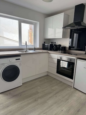 A modern kitchen with a washing machine, oven, microwave, and a window with a view of the outside. - Ground Floor Flat with Courtyard garden (Weston-super-Mare)