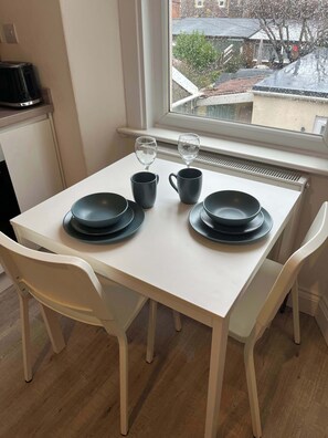 A dining table with black plates, two black cups, and two wine glasses, set in a room with white chairs and a window. - 1 Bed Room Flat Close to seafront (Weston-super-Mare)