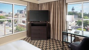 A hotel room with a bed, a TV, a desk, and large windows offering city views.