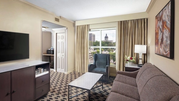 A hotel room with a flat-screen TV, a sofa, a chair, a small table, a window with curtains, and a view of a building.