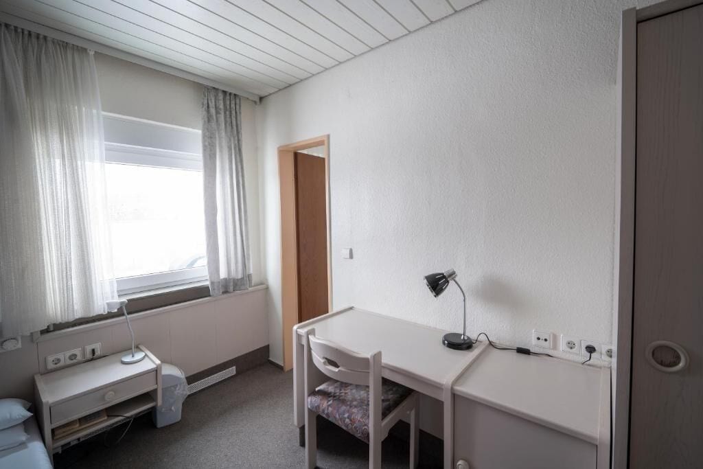 Ein kleiner, schlicht eingerichteter Raum mit Schreibtisch, Stuhl und Bett.