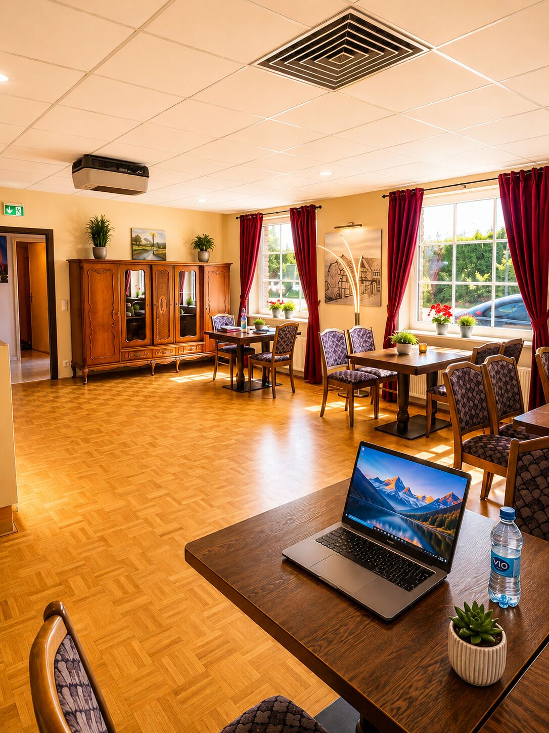 Een hotelkamer met houten vloer, een zithoek met rode gordijnen, een laptop op een tafel en een raam met uitzicht op groen.
