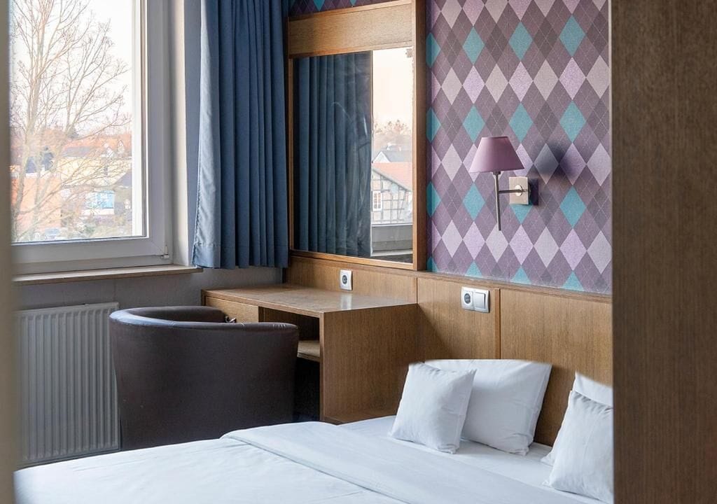 A hotel room with a bed, a desk, a chair, a window with curtains, and a wall with a diamond pattern.