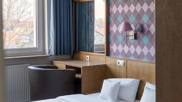 A hotel room with a bed, a desk, a chair, a window with curtains, and a wall with a diamond pattern.