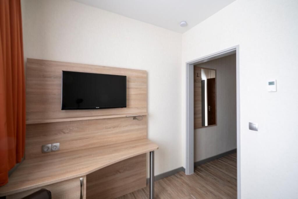 Habitación de hotel con televisor montado, escritorio con cajones, puerta que da a otra habitación e interruptor de luz en la pared.