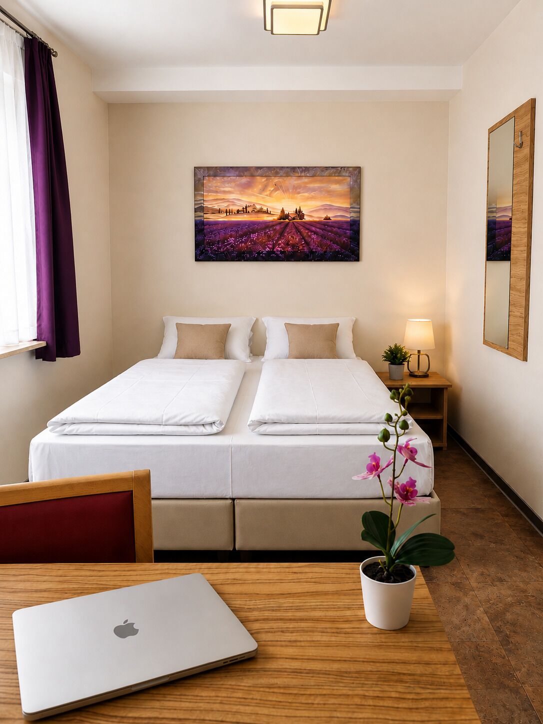 Habitación de hotel con cama, escritorio, ordenador portátil, mesita de noche con lámpara y una orquídea en maceta.