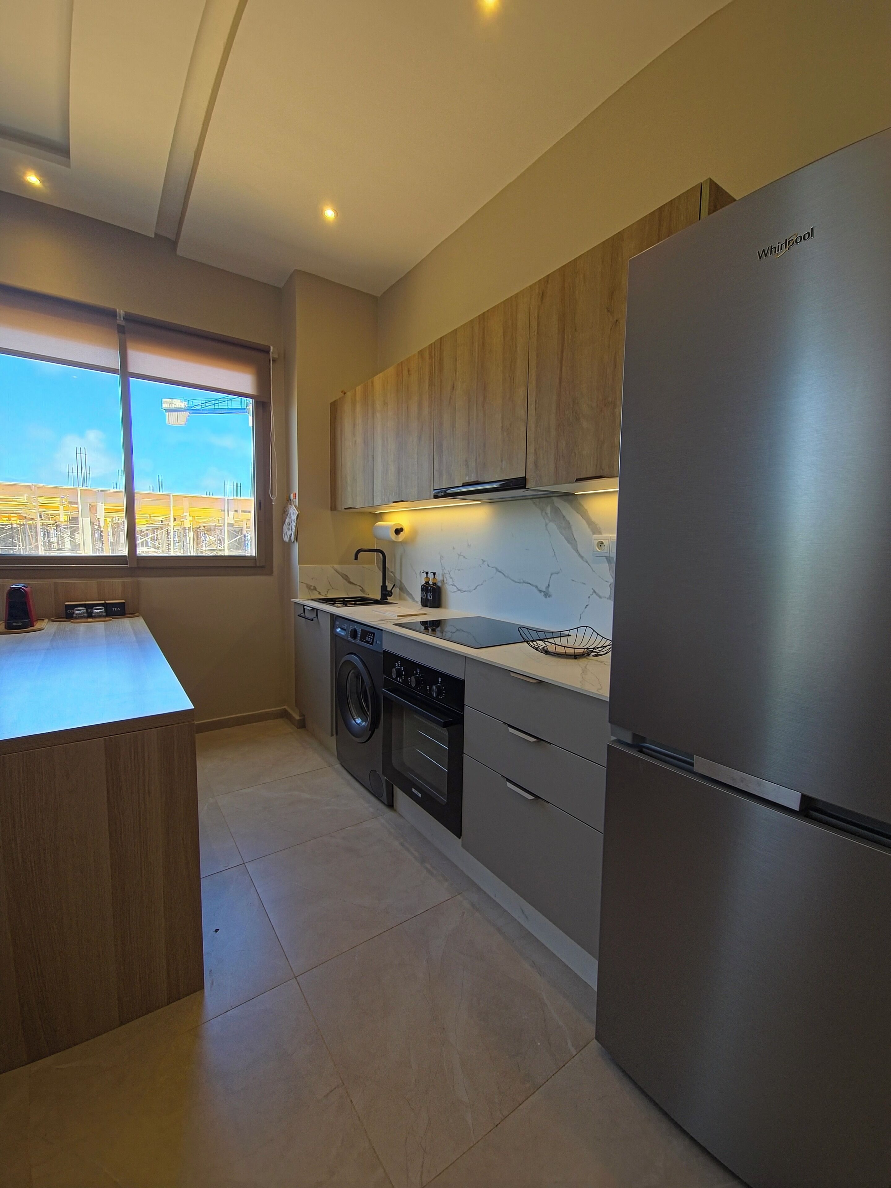 Una cocina moderna con un refrigerador de acero inoxidable, armarios de madera, lavadora integrada y una ventana con vistas a una obra en construcción.