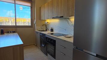Una cocina moderna con un refrigerador de acero inoxidable, armarios de madera, lavadora integrada y una ventana con vistas a una obra en construcción.