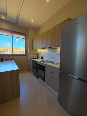 Una cocina moderna con un refrigerador de acero inoxidable, armarios de madera, lavadora integrada y una ventana con vistas a una obra en construcción.