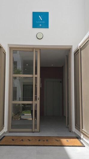 Entrada de hotel con puerta de vidrio, un cartel azul encima y felpudo en la entrada.