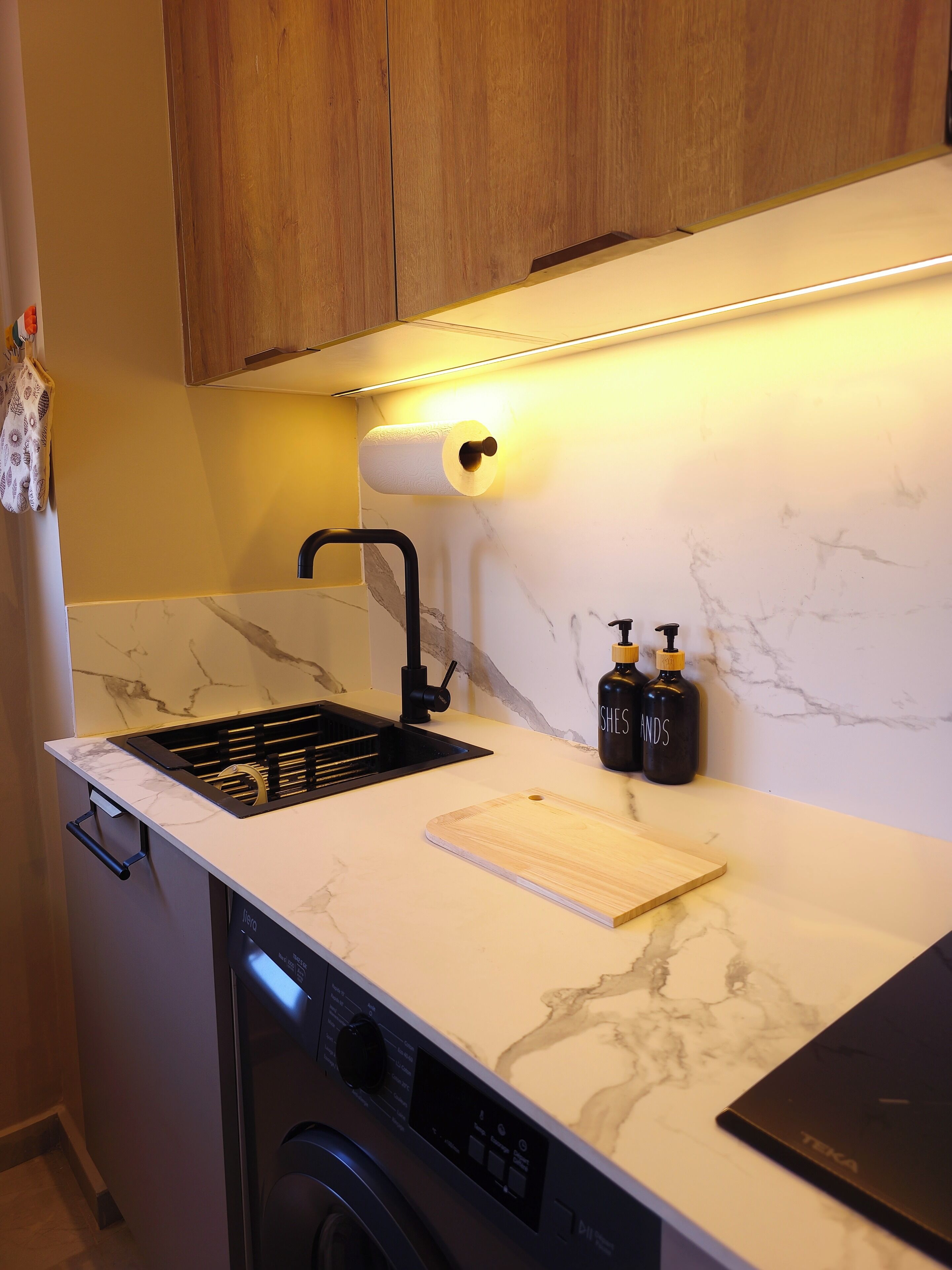 A compact kitchen with a built-in dishwasher, a sink with a black faucet, a marble countertop, and wooden cabinets.