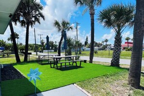 A grassy area with picnic tables and umbrellas, surrounded by palm trees.