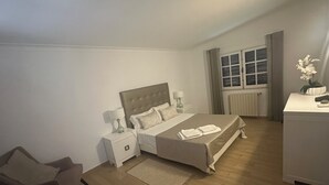 Ein Schlafzimmer mit Bett, Nachttischen, einem Sessel, einem Schreibtisch und einem Fenster mit Vorhängen.