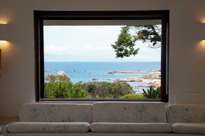 Ein Fenster mit Blick auf das Meer und die felsige Küste, ein Sofa mit einem gemusterten Kissen und eine Wandleuchte.