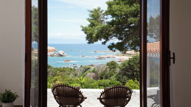 A view through a glass door to a balcony with two wicker chairs, a potted plant, and a coastal landscape with trees and the sea. - Domus Gallesi by Wonderful Italy (Portobello)