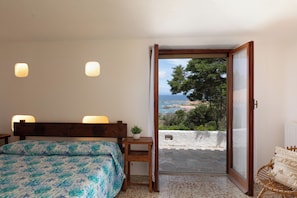 Ein Schlafzimmer mit Bett, Nachttischlampen, einem kleinen Tisch mit Pflanze und Meerblick durch eine offene Tür.