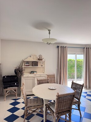 Zona de dinar amb taula rodona, cadires de vimetla, un armari blanc amb objectes decoratius i una vista per una porta de vidre.