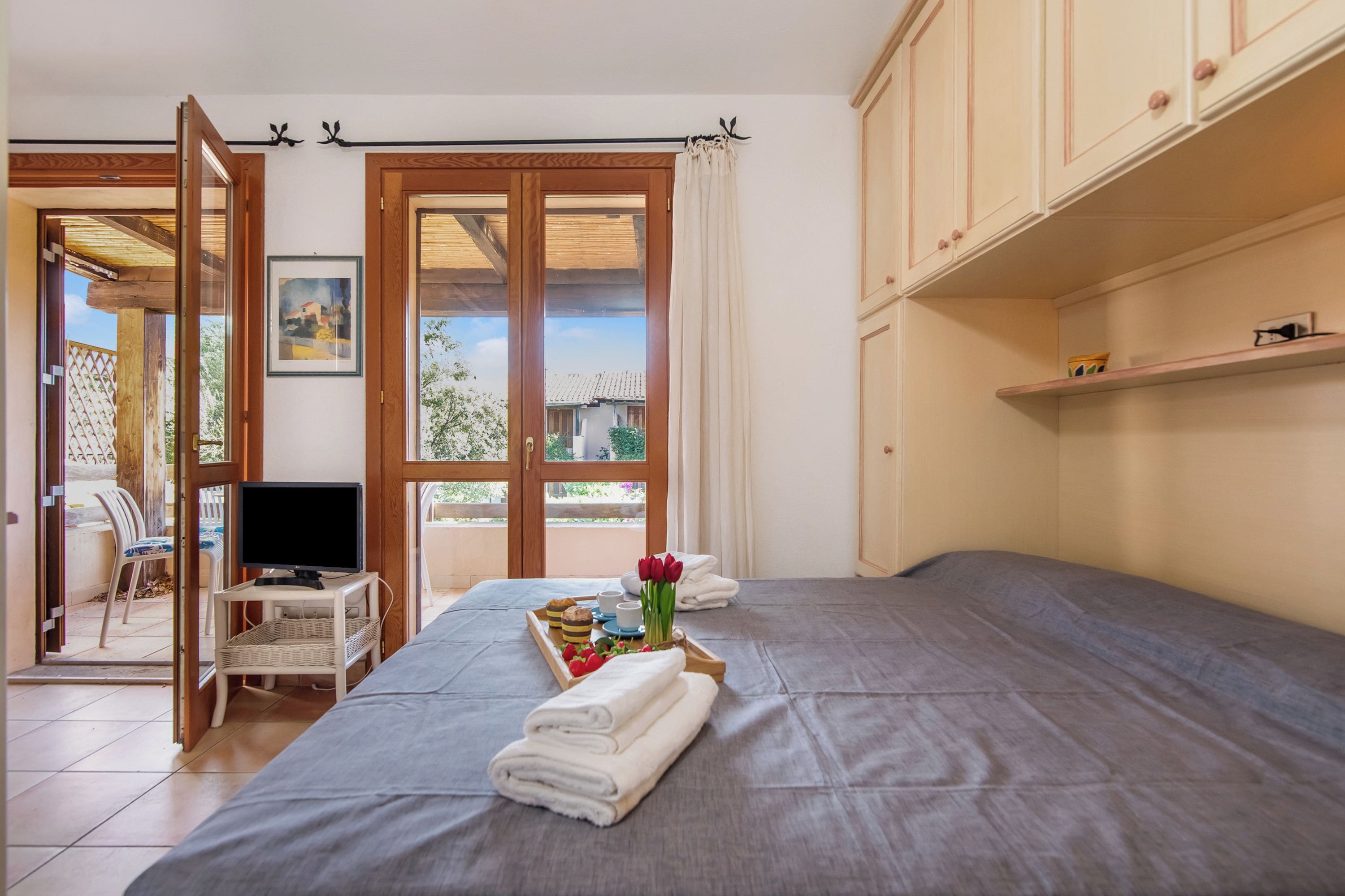 A bedroom with a bed, a television on a stand, a tray with towels and flowers, and a view through a door.
