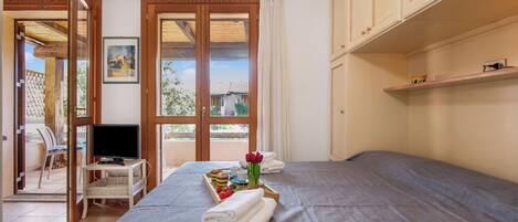 A bedroom with a bed, a television on a stand, a tray with towels and flowers, and a view through a door.