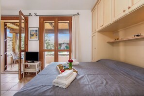 Quarto com cama, televisão sobre suporte, bandeja com toalhas e flores, e vista através de uma porta.