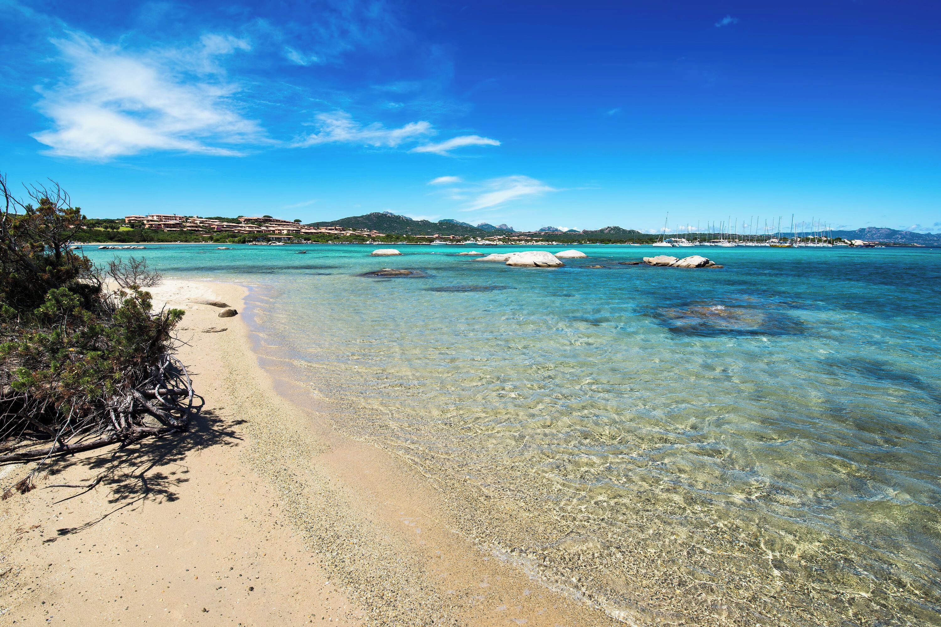 Une plage à l’eau turquoise et limpide, un rivage sablonneux et une vue lointaine sur une ville côtière avec ses bâtiments.