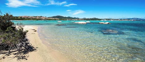 Une plage à l’eau turquoise et limpide, un rivage sablonneux et une vue lointaine sur une ville côtière avec ses bâtiments.