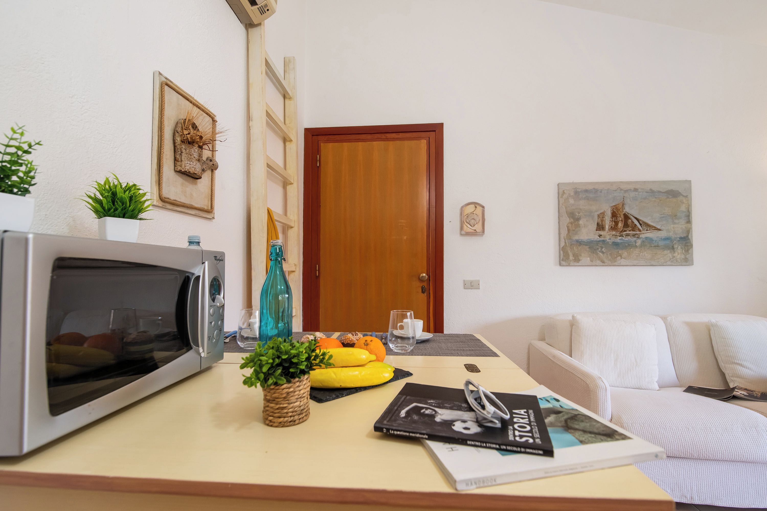 A kitchen with a microwave, a wooden door, a white sofa, and a painting on the wall.
