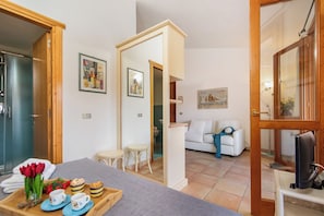 A room with a bed, a TV, a small table with a tray of food, and a white sofa. - Nido Di Tom (Olbia)