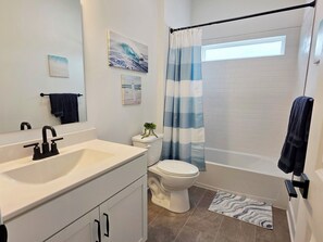 Een badkamer met een witte wastafel, toilet en badkuip. Er is een handdoekhouder, een kamerplant en een raam met gordijn.