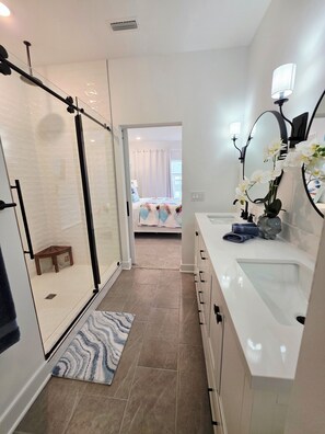 A modern bathroom with a glass shower enclosure, a white sink with a mirror, and a view into the bedroom. - Brand New Spacious 4-bedroom townhome with pool and gym. (Selbyville)