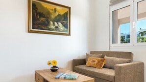 A living room with a sofa, a coffee table with a book and vase, a painting on the wall, and a window with a view. - Ocean Alley Seaside Retreats (K. Gouves)