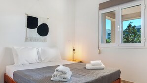 A hotel room with a bed, pillows, a wall hanging, a bedside lamp, a window, and a view of greenery outside. - Ocean Alley Seaside Retreats (K. Gouves)
