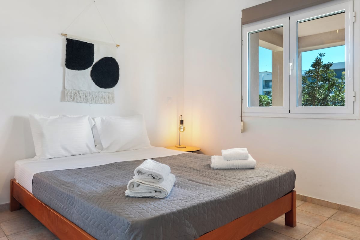 Een hotelkamer met een bed, kussens, een wanddecoratie, een bedlampje, een raam en uitzicht op groen.