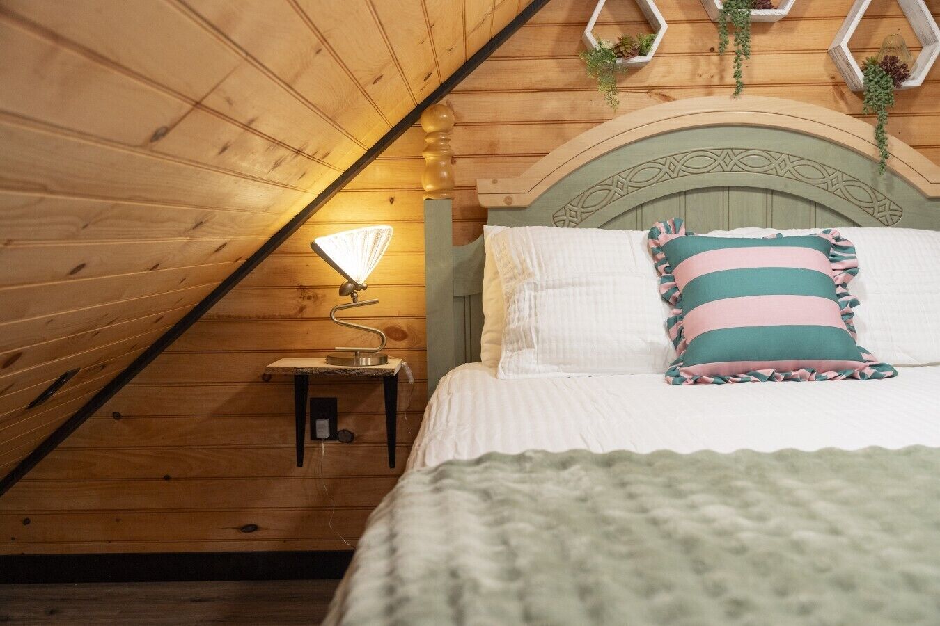 Un dormitorio acogedor con techo inclinado, cabecero de madera, lámpara de noche y almohada a rayas.