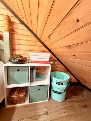 Una esquina de una habitación con techo de madera inclinado, un estante blanco con cajas de almacenamiento, una caja de Scrabble, una cesta y un cubo de basura azul.