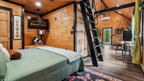 A wooden cabin interior with a bed, a ladder, a desk, and a sign that reads 'StayOver.' - The Grey Squirrel Hideaway - Hot Tub , Fire-Pit, Pet-Friendly (Campton)