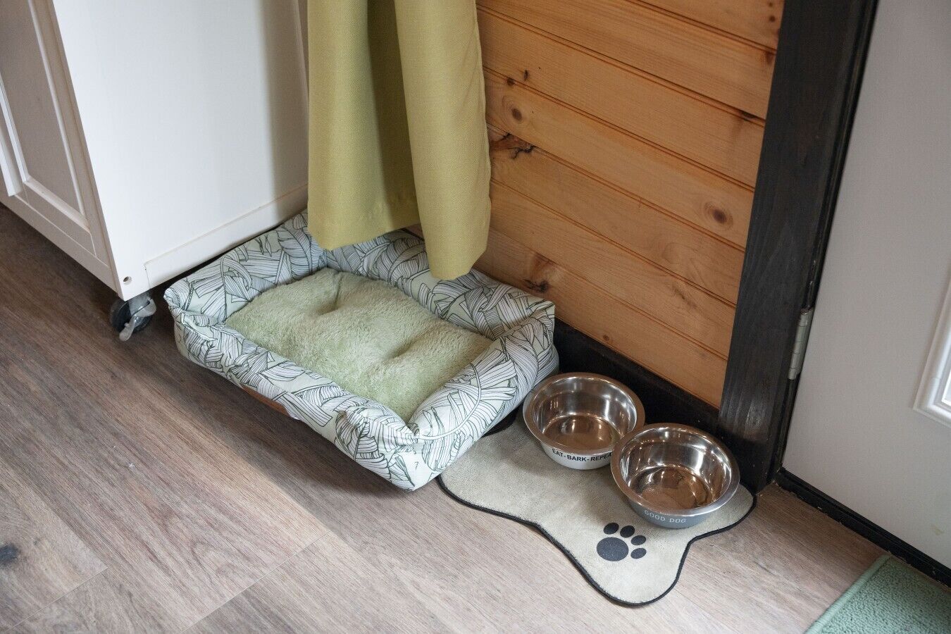 A pet bed with a patterned edge, two metal bowls, and a paw print mat on a wooden floor.
