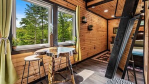 A wooden interior with a table and stools, large windows, and a view of trees. - The Grey Squirrel Hideaway - Hot Tub , Fire-Pit, Pet-Friendly (Campton)