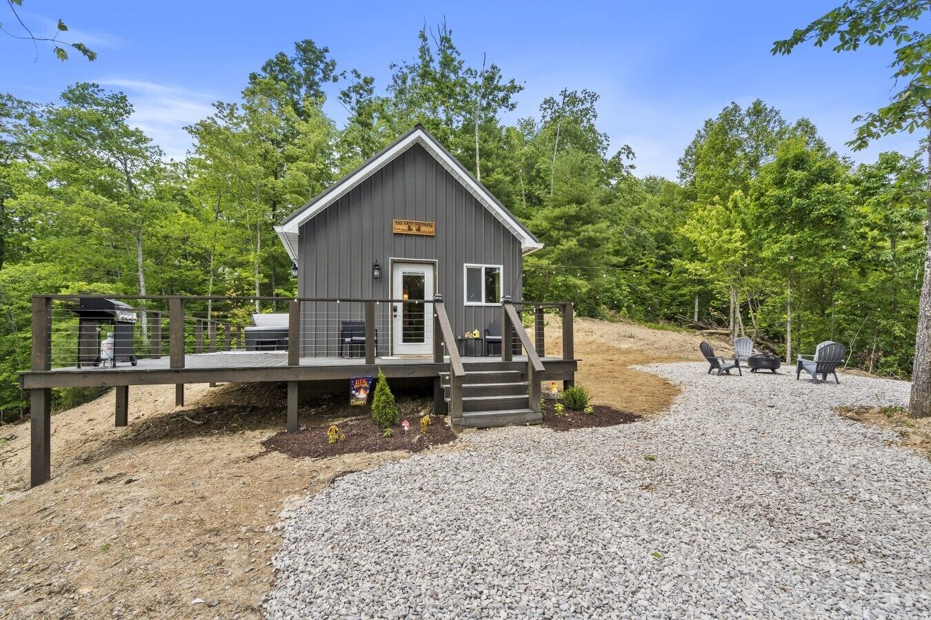 A small cabin with a deck, surrounded by trees and gravel.